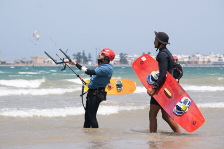 Kitesurf Student heading ou two kite with kite instructor holding the kite board in front of UNESCO old Medina and port of Essaouira Morocco.